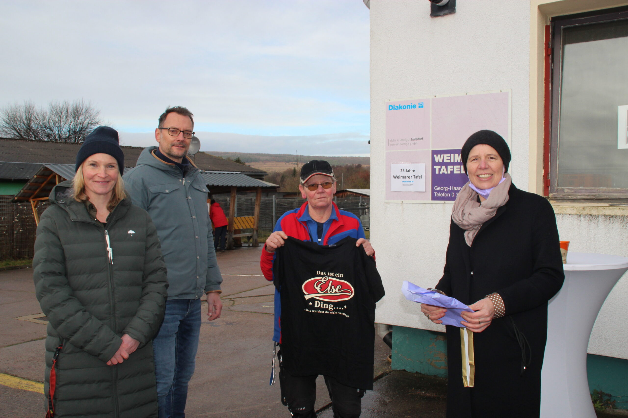 Sozialkontor Johannes Falk, Tafel Weimar, vier Personen am Hausschild, eine Frau zeigt ein T-shirt