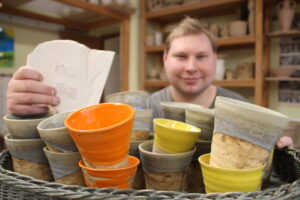Töpferei Werkstätten Christopheruhof Altengesees, junger Mann vor farbigen Saftbechern aus Ton