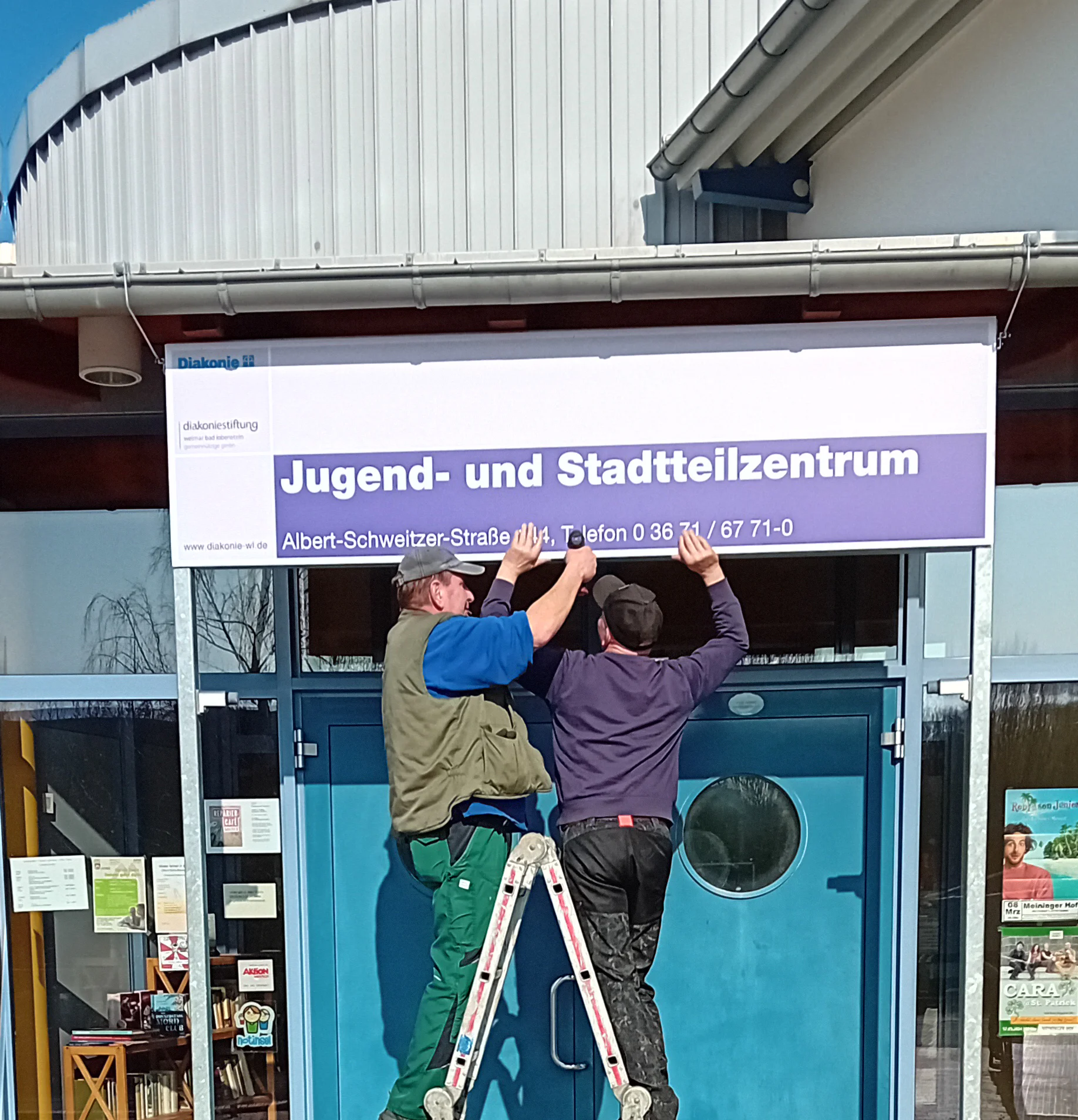 zwei Männer bringen das Eingangsschild zum Jugend- und Stadtteilzentrum Saalfeld an