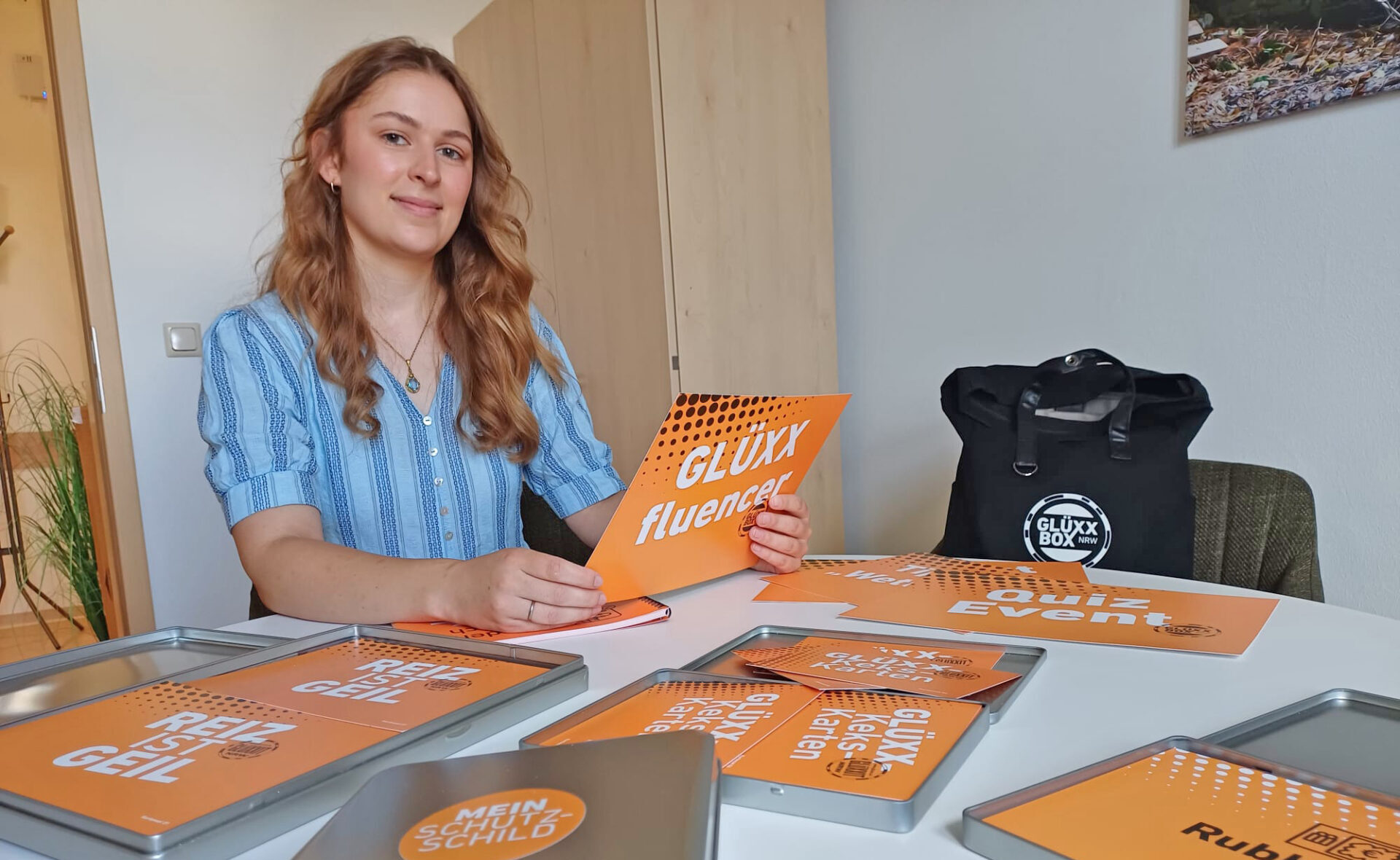 Suchrberatung-eine Mitarbeiterin zeigt den Inhalt der Glüxxbox, Alukoffer mit orangefarbenen Themenkarten